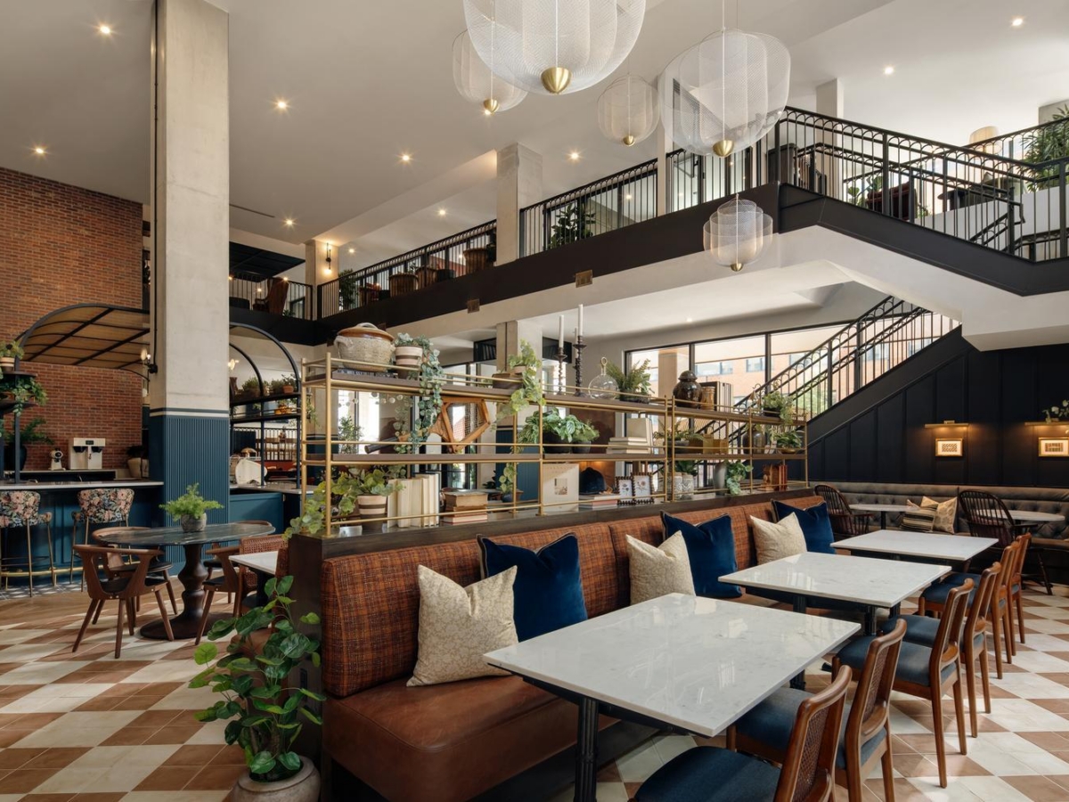 Modern, spacious café with high ceilings, large windows, and stylish decor. Features marble tables, cushioned seating, potted plants, and a two-level layout with metal railings and warm ambient lighting.