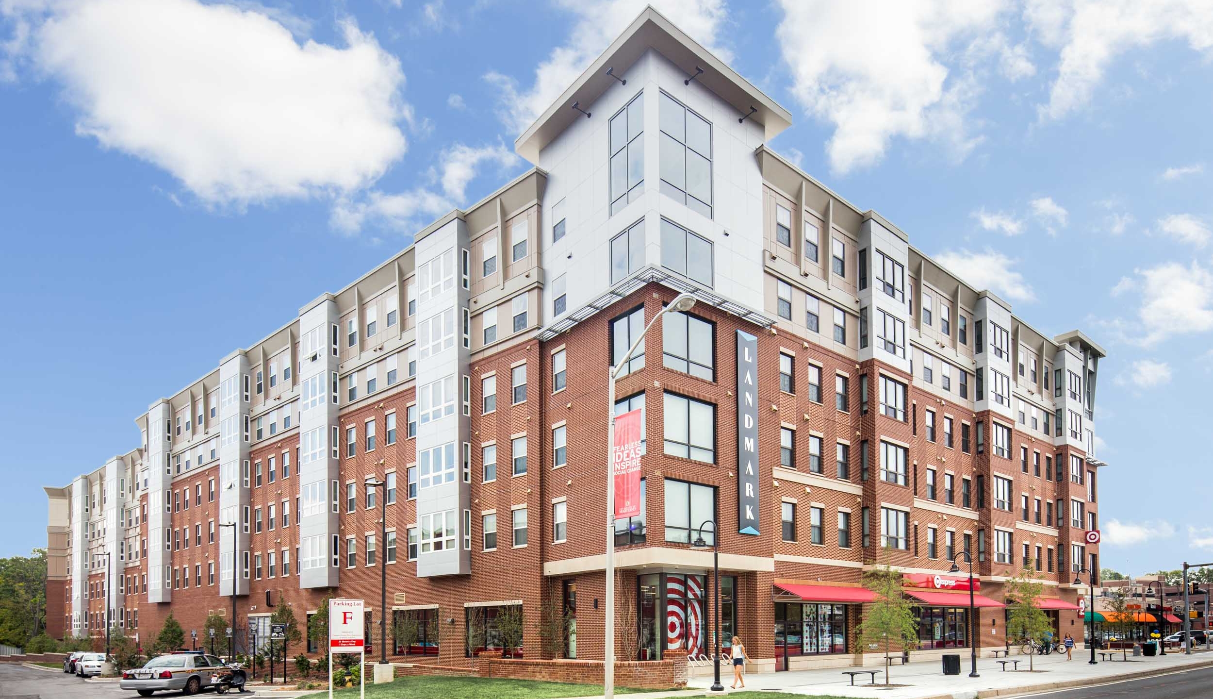 A modern, multi-story apartment building with ground-level retail stores, including a Target. Cars are parked along the street, and the sky is blue with scattered clouds.