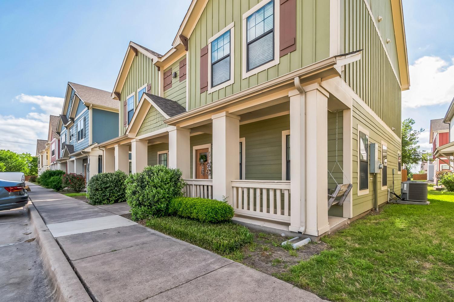 Green two-story house with a covered front porch, white trim, and brown shutters, located on a sidewalk-lined street with neighboring homes and a parked car. Bushes and grass border the house.