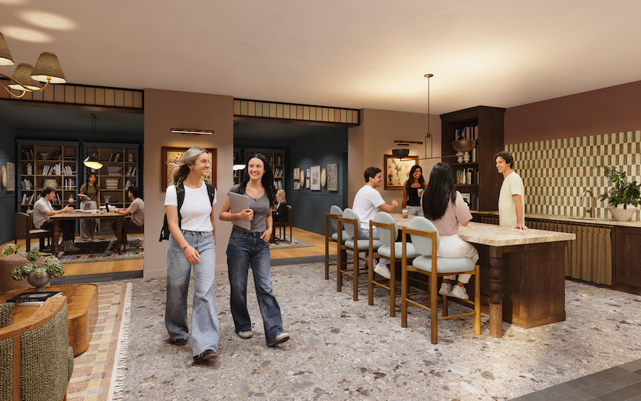 People socialize in a modern, open lounge at Rambler Blacksburg student housing, surrounded by bookshelves, a kitchen island, and relaxed seating. Two women walk and chat while others sit, talk, and read in a cozy, warmly lit environment.