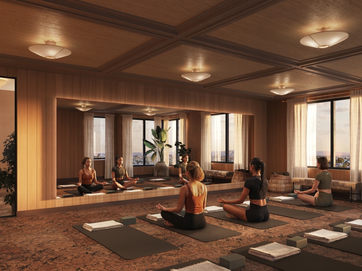Five people sit on yoga mats in a serene, sunlit studio with wooden walls and large mirrors at Rambler Blacksburg student housing. One person enters the room, and the windows reveal a cityscape outside. Towels and blocks are neatly placed by each mat.
