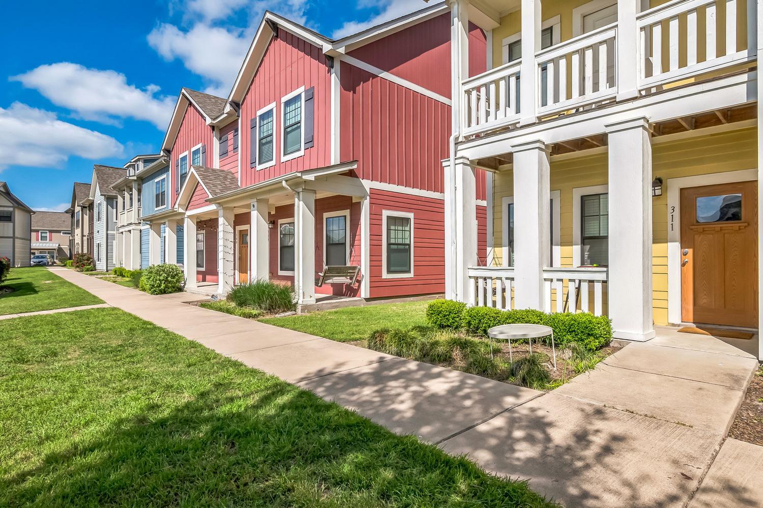 Row of colorful modern townhouses with porches, large windows, and neatly trimmed lawns on a sunny day with blue sky and scattered clouds.