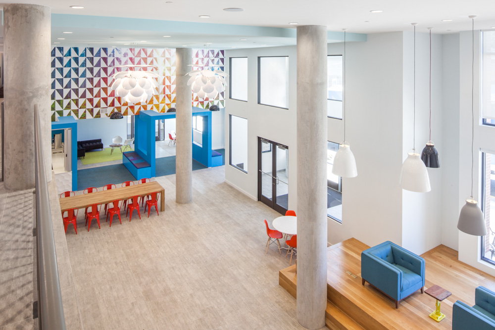 A bright, modern open space with tall ceilings, large windows, colorful geometric wall art, long dining table with red chairs, small lounge areas, and hanging pendant lights. Wooden steps lead to a blue sofa and chairs.