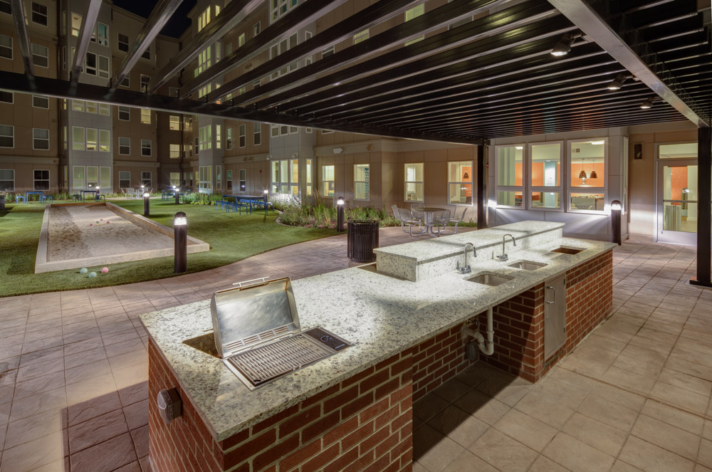 Outdoor communal kitchen area with a stainless steel grill and sinks set in a granite countertop, under a pergola. Apartment building and grassy courtyard with bocce court are visible in the background at night.