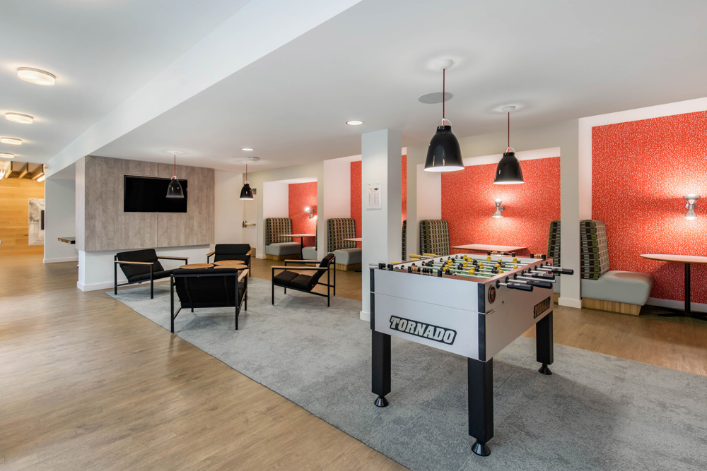 Modern lounge area with a foosball table in the center, surrounded by black chairs, a wall-mounted TV, and booth seating with red patterned walls and pendant lights. The space has wood floors and a cozy, open layout.