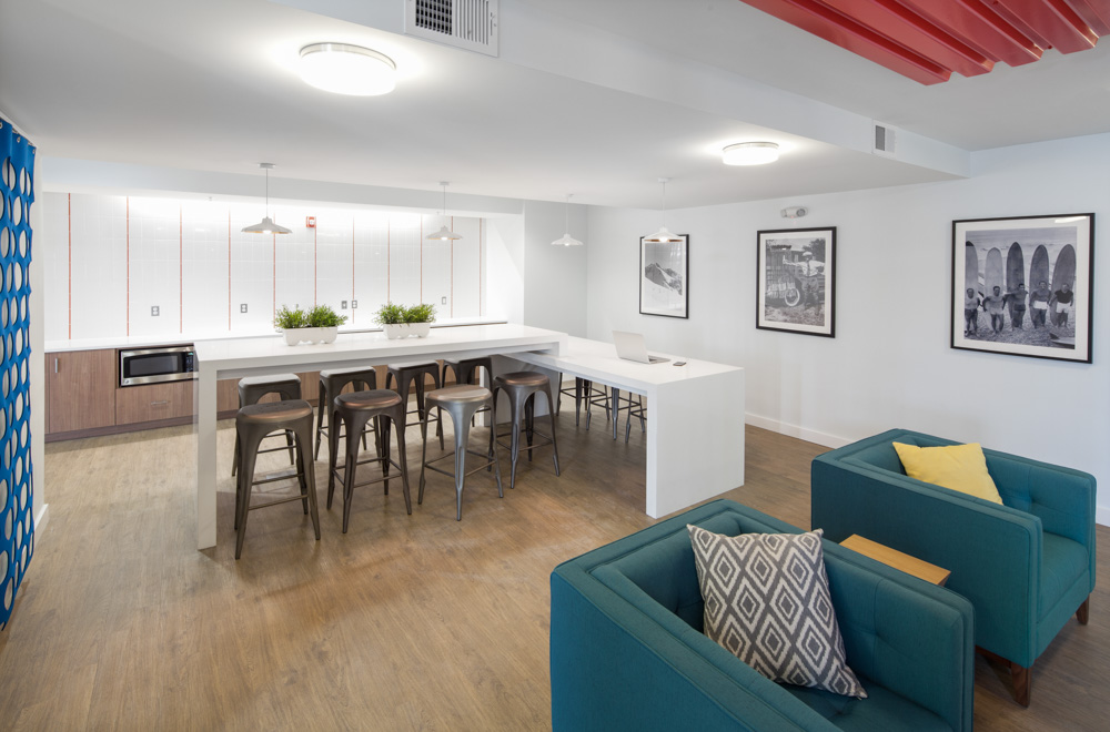 Modern communal kitchen and lounge area with a white island, stools, green sofas with pillows, framed wall art, potted plants, wood flooring, and bright overhead lighting.