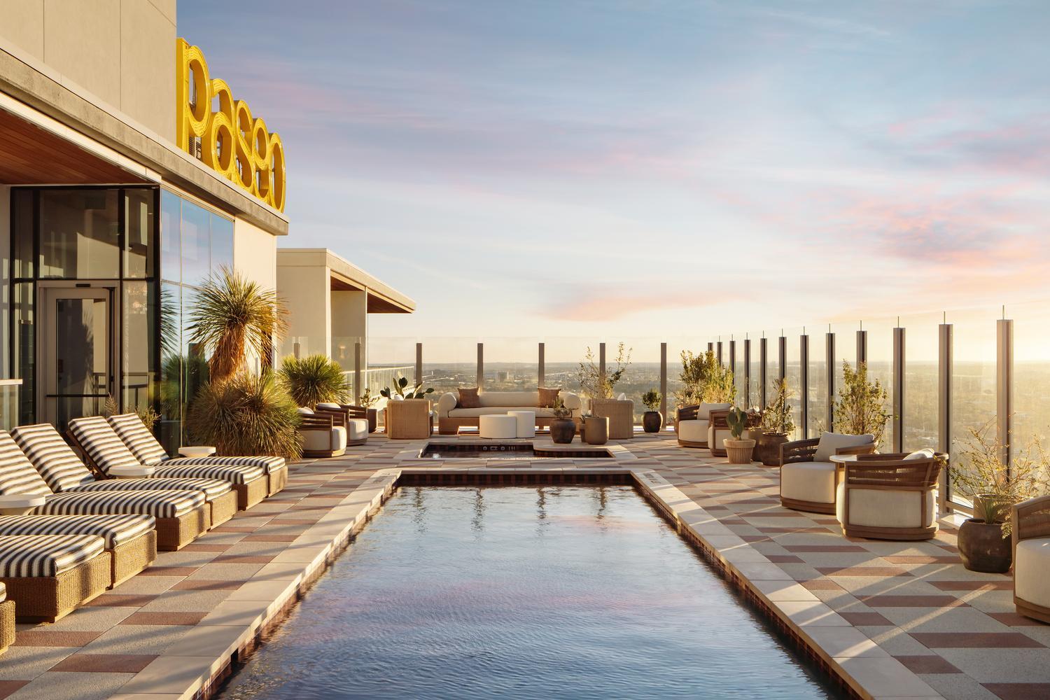 Rooftop pool area with striped lounge chairs, modern seating, glass safety panels, and plants. A large yellow sign is on the building. The city skyline and a colorful sunset are visible in the background.