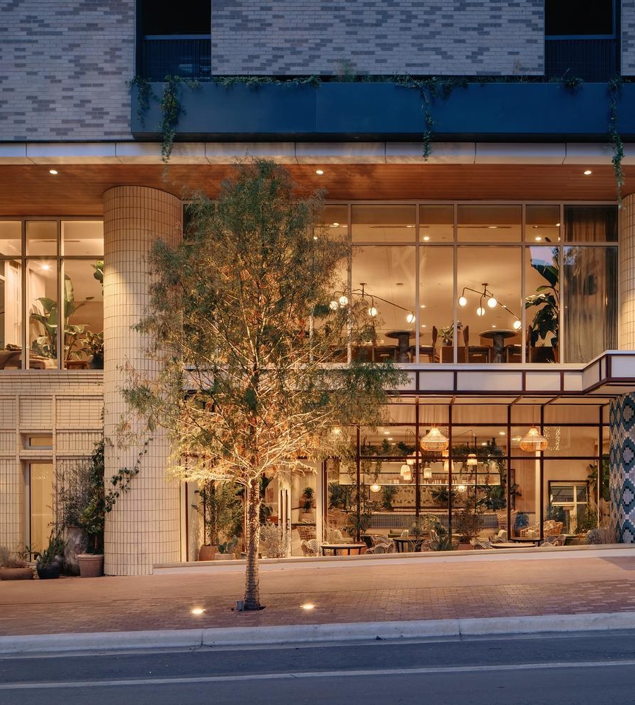 Street view of a modern coffee shop with large glass windows, warm lighting, potted plants, and trees outside; upper level is visible with seating and hanging lights. COFFEE is written on a white exterior wall.