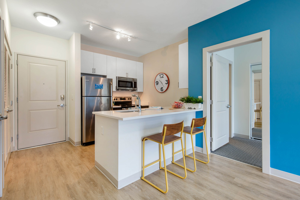 Modern kitchen with white cabinets, stainless steel appliances, a large clock on the wall, and a blue accent wall. A white island counter has two wooden barstools with yellow legs. Wood flooring and bright lighting.