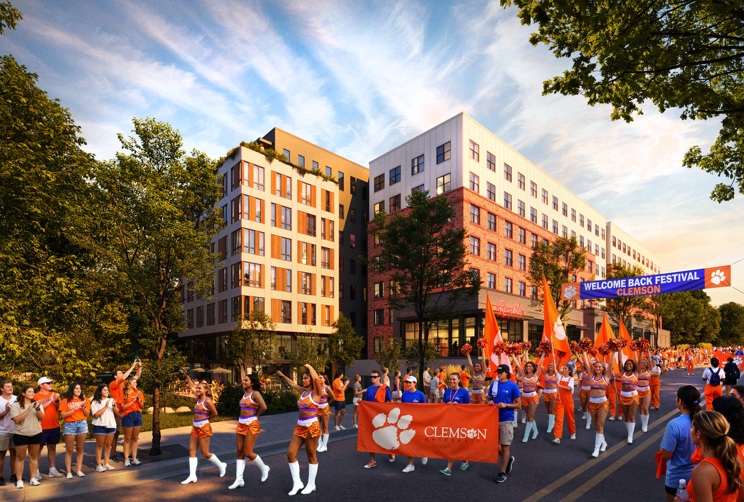 A parade of cheerleaders and participants in orange outfits carrying a Clemson banner marches down a street lined with people, with modern buildings and trees in the background under a blue sky. A Welcome Back Festival sign is overhead.