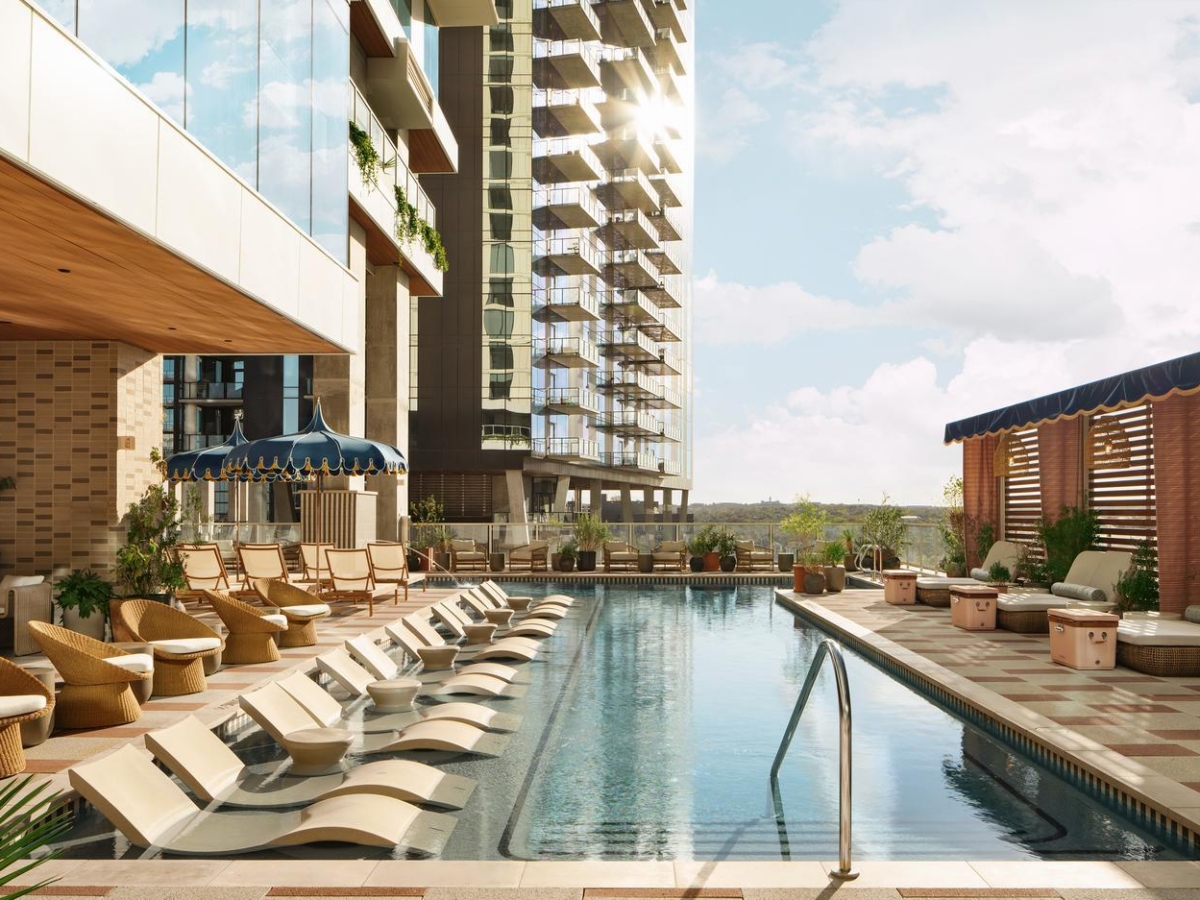 A modern outdoor pool area with lounge chairs partially immersed in water, shaded seating, potted plants, and city buildings in the background under a sunny sky.