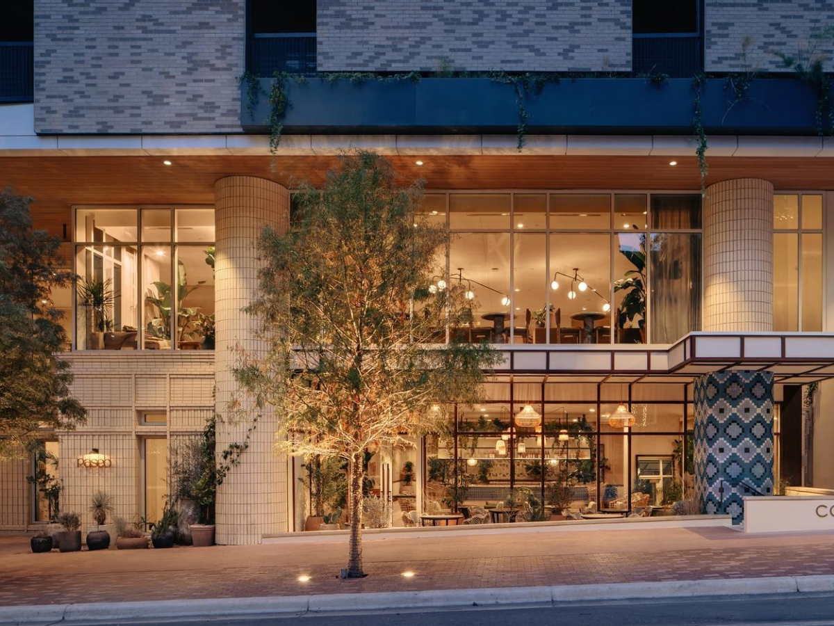 A modern coffee shop with large glass windows, warm lighting, and greenery on the sidewalk and balcony; the building features tile accents and a sign that says COFFEE near the entrance.