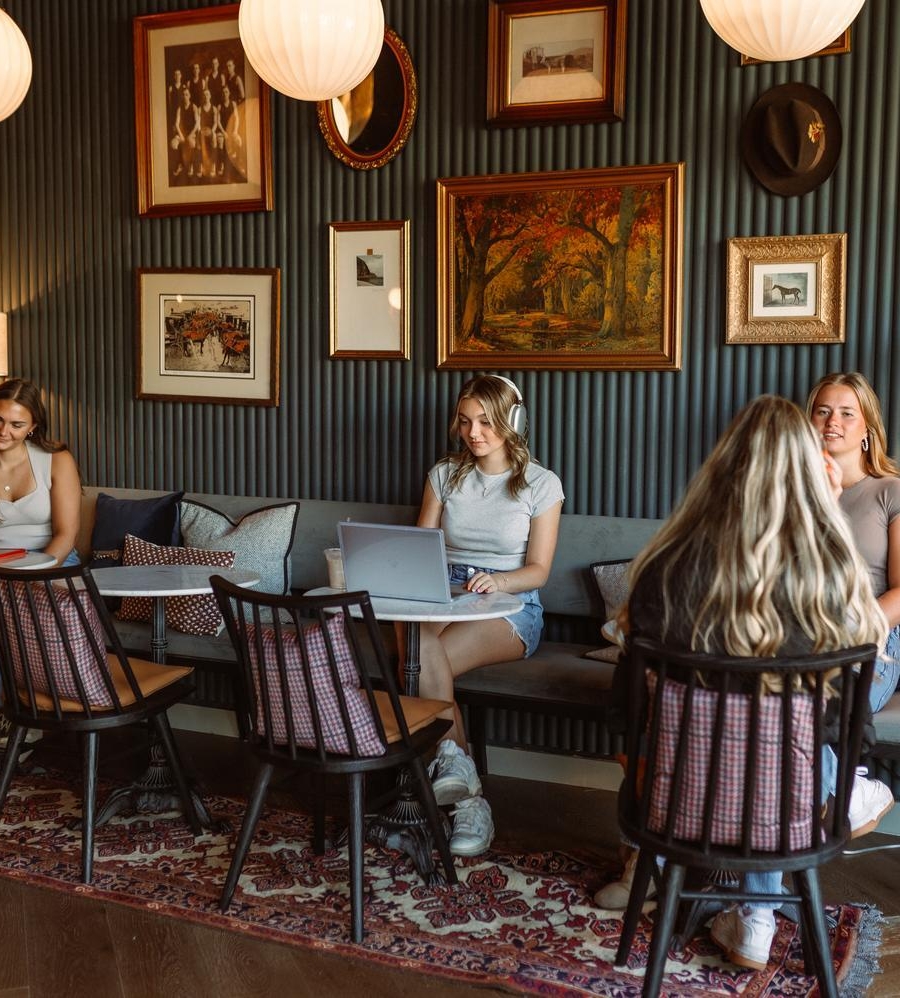 Five young women sit and chat around two small tables in a cozy, warmly lit room with framed art on the walls, a patterned rug, and plants. One uses a laptop while others talk and use their phones.