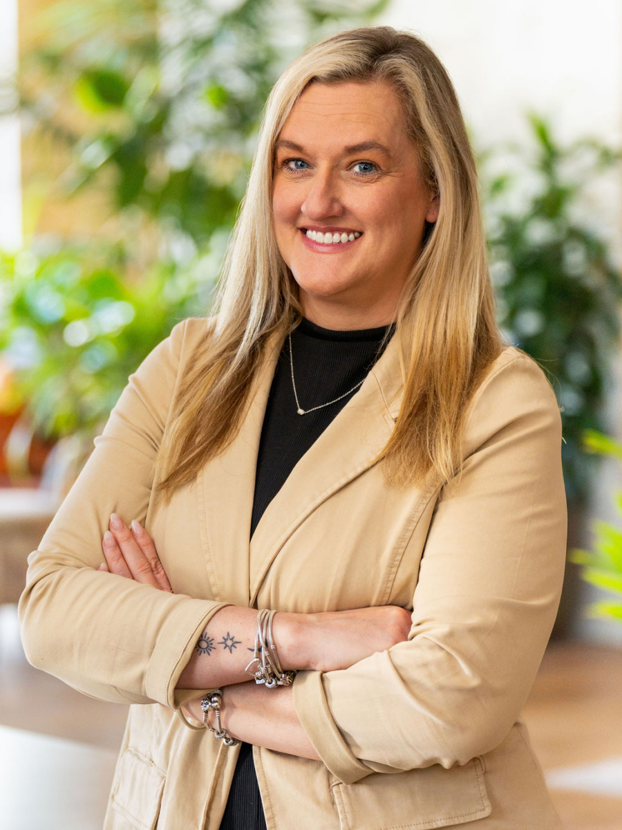 A woman with long blonde hair wearing a beige blazer and black top stands with arms crossed, smiling in a bright indoor setting with green plants in the background.