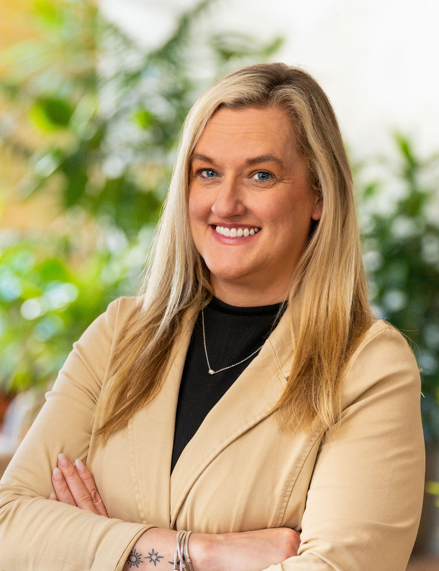 A woman with long blonde hair wearing a beige blazer and black top stands with arms crossed, smiling in a bright indoor setting with green plants in the background.