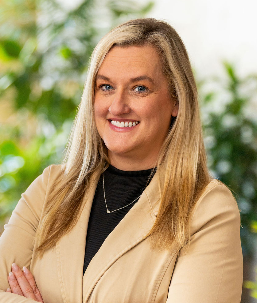 A woman with long blonde hair wearing a beige blazer and black top stands with arms crossed, smiling in a bright indoor setting with green plants in the background.