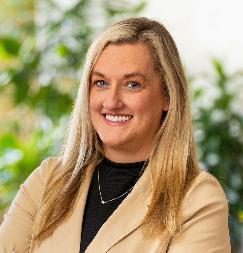 A woman with long blonde hair wearing a beige blazer and black top stands with arms crossed, smiling in a bright indoor setting with green plants in the background.