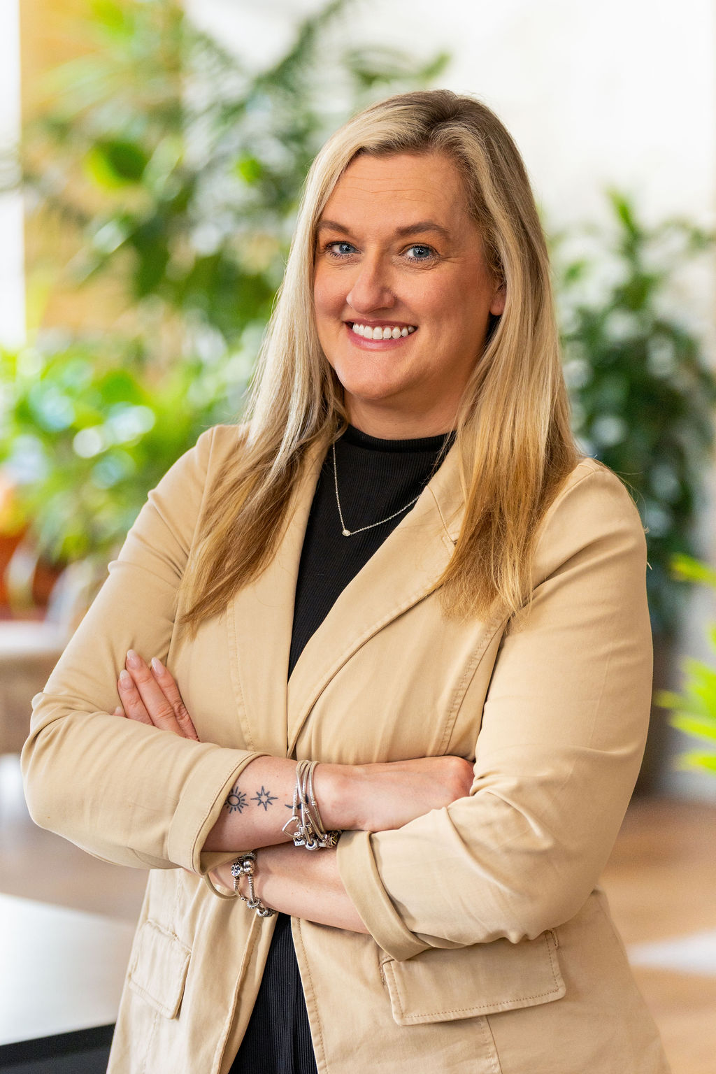 A woman with long blonde hair wearing a beige blazer and black top stands with arms crossed, smiling in a bright indoor setting with green plants in the background.