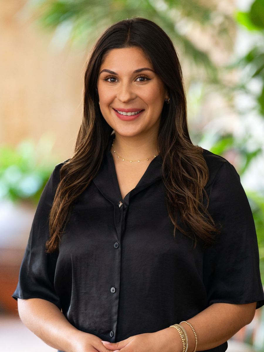 A woman with long brown hair and a black blouse smiles while standing indoors. She wears gold jewelry and is surrounded by a blurred background with green plants.