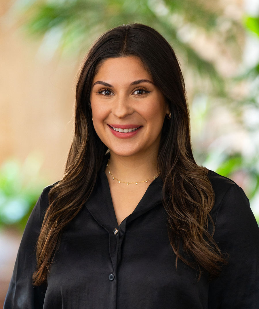 A woman with long brown hair and a black blouse smiles while standing indoors. She wears gold jewelry and is surrounded by a blurred background with green plants.