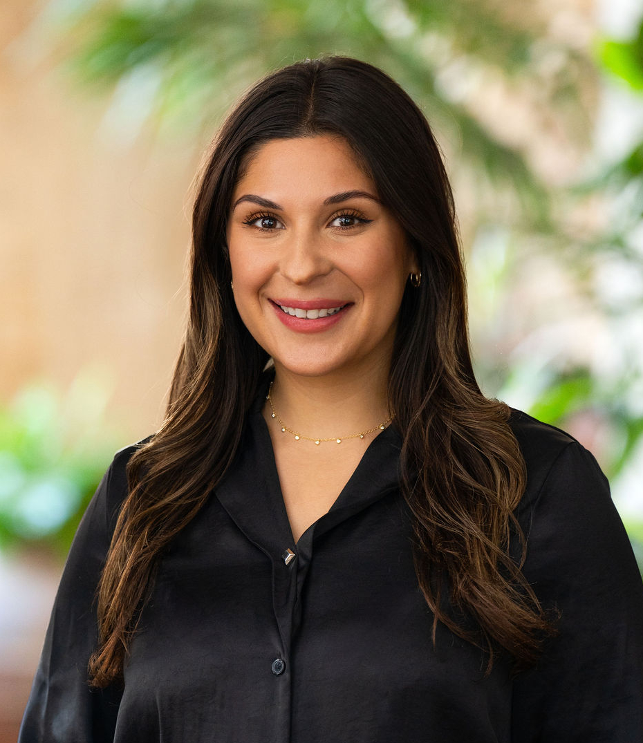 A woman with long brown hair and a black blouse smiles while standing indoors. She wears gold jewelry and is surrounded by a blurred background with green plants.