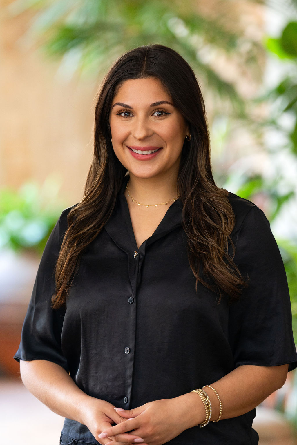 A woman with long brown hair and a black blouse smiles while standing indoors. She wears gold jewelry and is surrounded by a blurred background with green plants.