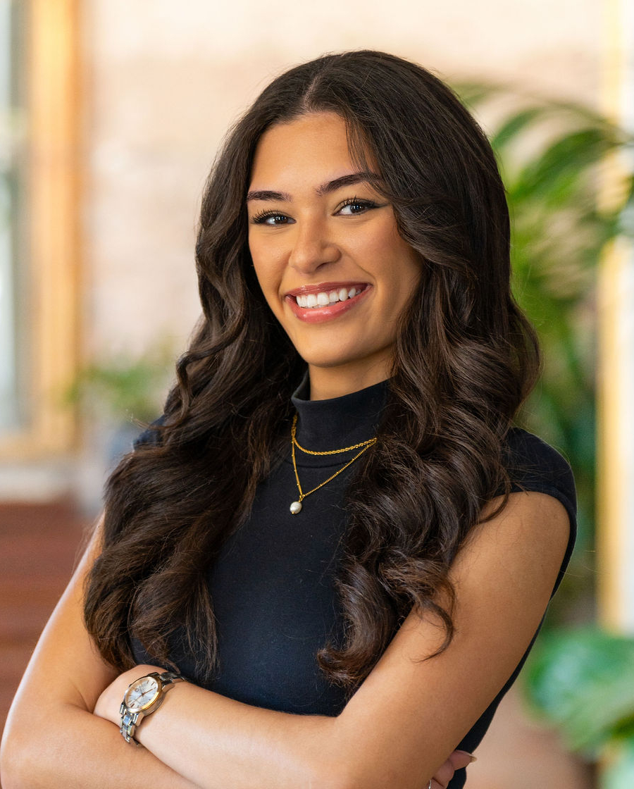 A woman with long, wavy dark hair stands smiling with arms crossed, wearing a black sleeveless top, layered gold necklaces, a watch, and blue jeans. The background features blurred greenery and warm indoor lighting.