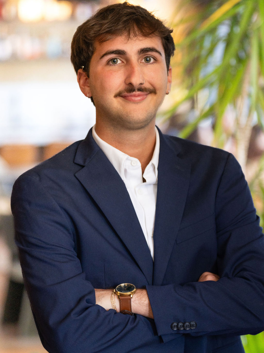 A young man with short brown hair and a mustache, wearing a navy blue suit and white shirt, stands with arms crossed, smiling slightly. The background is blurred with green plants and warm indoor lighting.