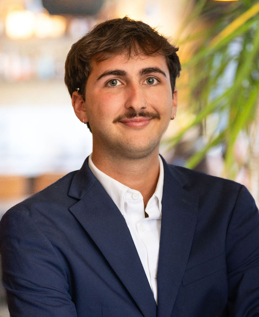 A young man with short brown hair and a mustache, wearing a navy blue suit and white shirt, stands with arms crossed, smiling slightly. The background is blurred with green plants and warm indoor lighting.
