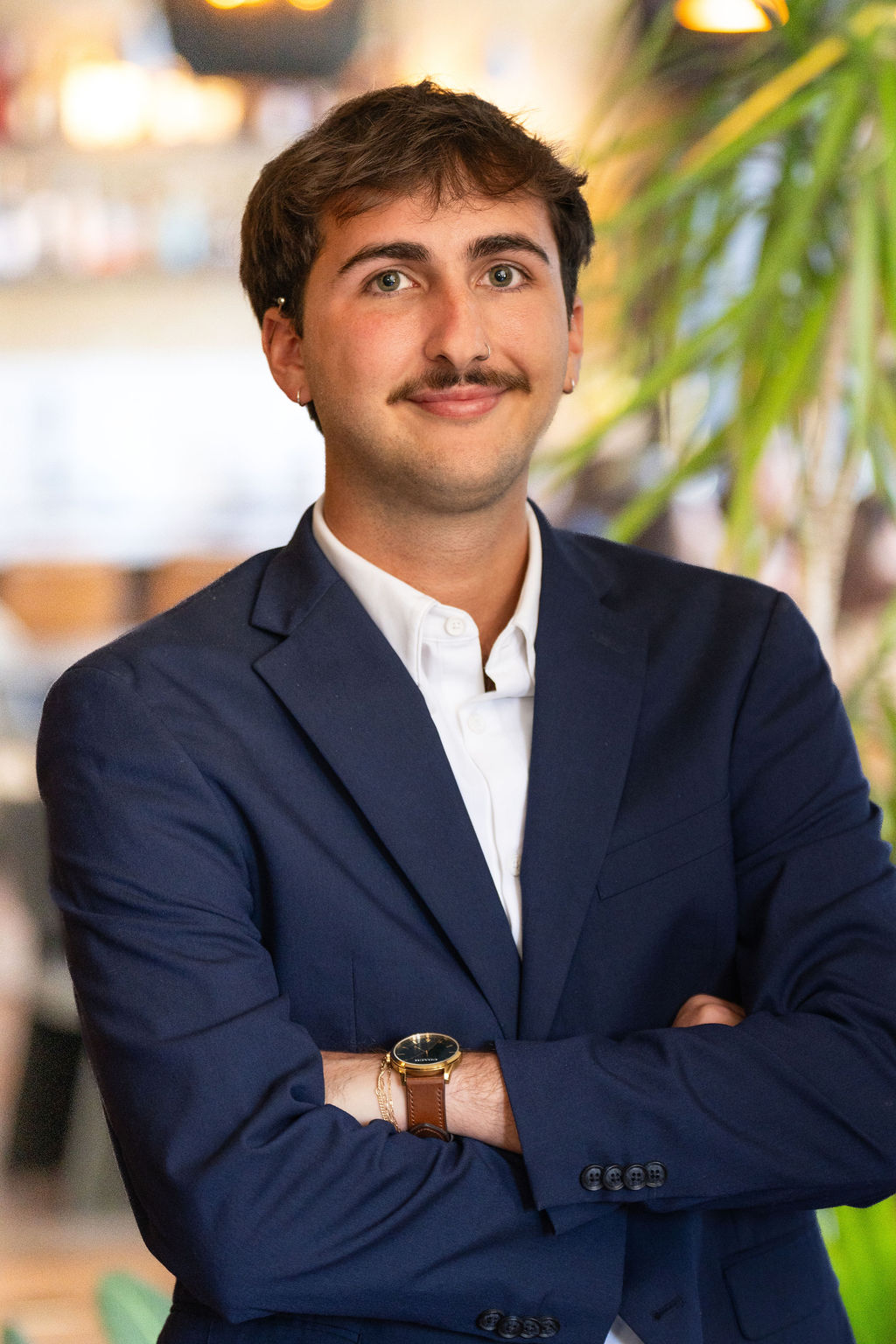 A young man with short brown hair and a mustache, wearing a navy blue suit and white shirt, stands with arms crossed, smiling slightly. The background is blurred with green plants and warm indoor lighting.