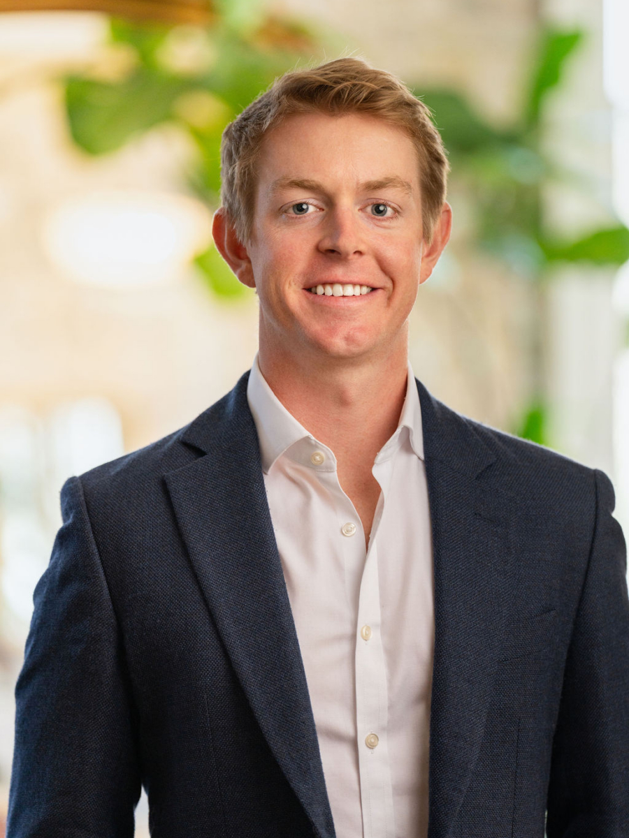 A young man with short blond hair, wearing a dark blue suit jacket over a white dress shirt, smiling and standing indoors with blurred green plants and light in the background.