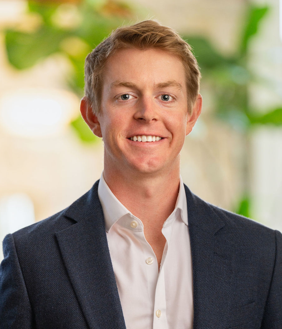 A young man with short blond hair, wearing a dark blue suit jacket over a white dress shirt, smiling and standing indoors with blurred green plants and light in the background.