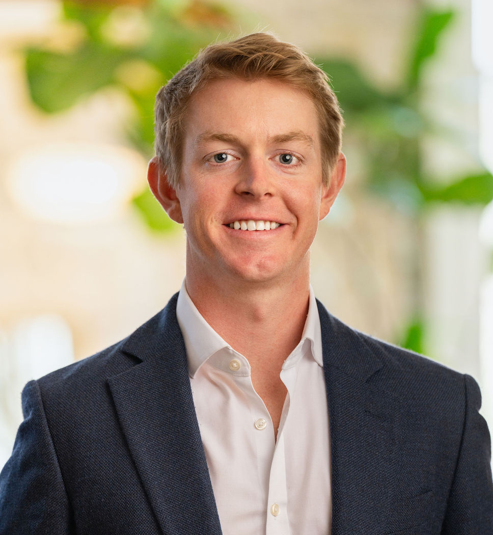 A young man with short blond hair, wearing a dark blue suit jacket over a white dress shirt, smiling and standing indoors with blurred green plants and light in the background.