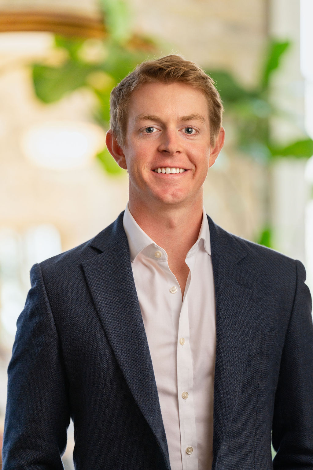 A young man with short blond hair, wearing a dark blue suit jacket over a white dress shirt, smiling and standing indoors with blurred green plants and light in the background.