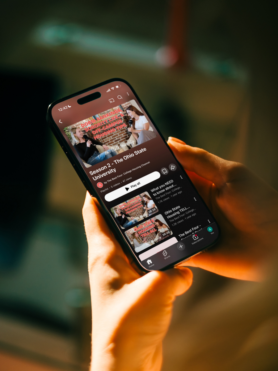 A person holds a smartphone displaying a YouTube video titled Сошники + Три Оси 2023 with related video thumbnails visible, against a blurred background.