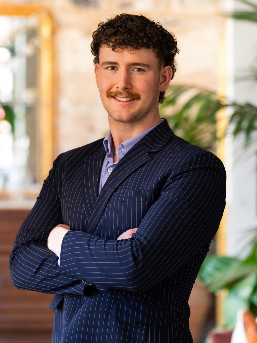 A man with curly brown hair and a mustache stands with arms crossed, wearing a navy pinstripe suit and purple shirt, smiling confidently indoors with plants and a gold-framed mirror in the background.