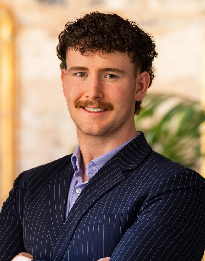 A man with curly brown hair and a mustache stands with arms crossed, wearing a navy pinstripe suit and purple shirt, smiling confidently indoors with plants and a gold-framed mirror in the background.