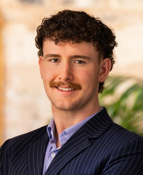 A man with curly brown hair and a mustache stands with arms crossed, wearing a navy pinstripe suit and purple shirt, smiling confidently indoors with plants and a gold-framed mirror in the background.