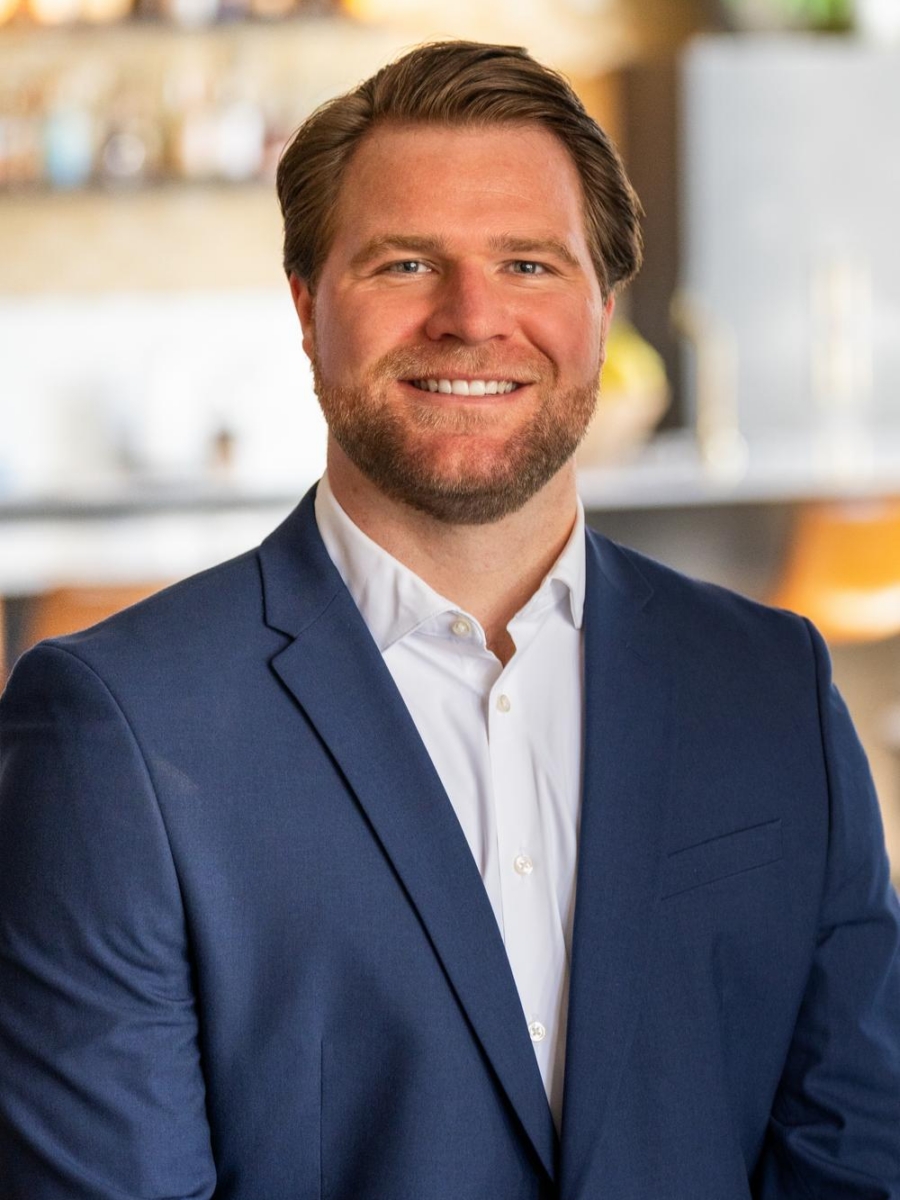 A man with short brown hair and a beard, wearing a blue suit and white shirt, smiles while standing indoors in a modern, softly blurred setting.