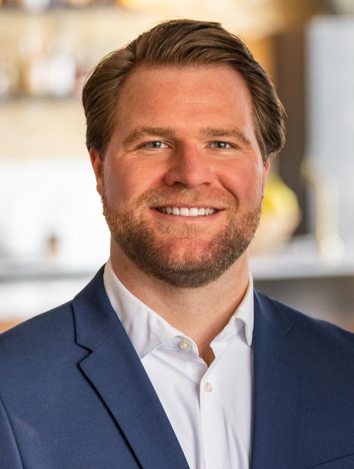 A man with short brown hair and a beard, wearing a blue suit and white shirt, smiles while standing indoors in a modern, softly blurred setting.