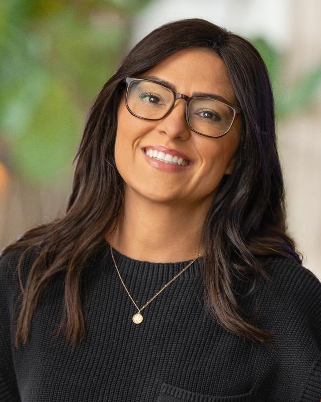 A woman with long dark hair and glasses smiles at the camera. She is wearing a black short-sleeved knit top with a front pocket, a gold necklace, and stands indoors with blurred greenery in the background.