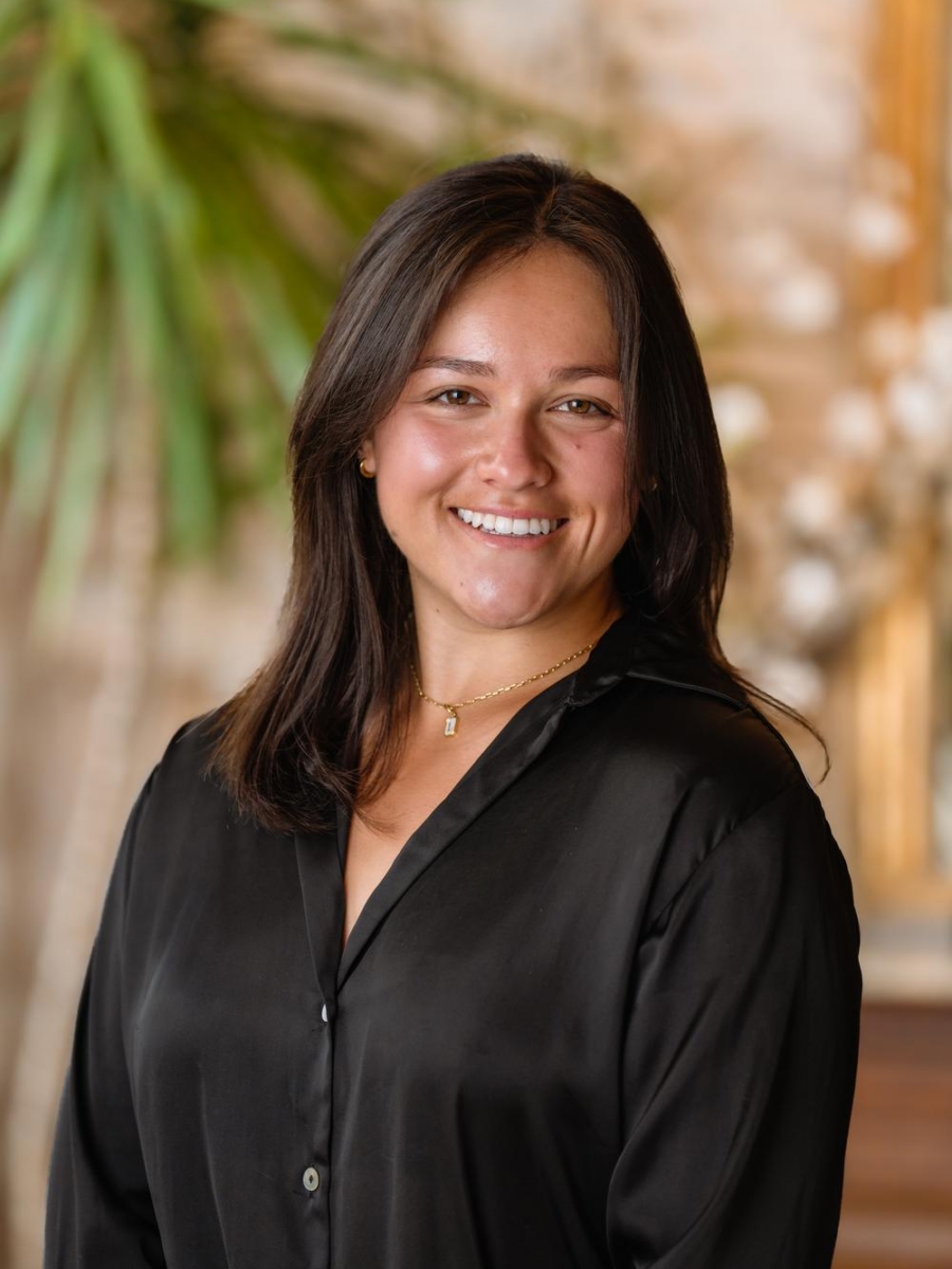 A woman with shoulder-length dark hair smiles while wearing a black button-up shirt and a gold necklace. She stands indoors with a blurred background featuring green plants and soft lighting.