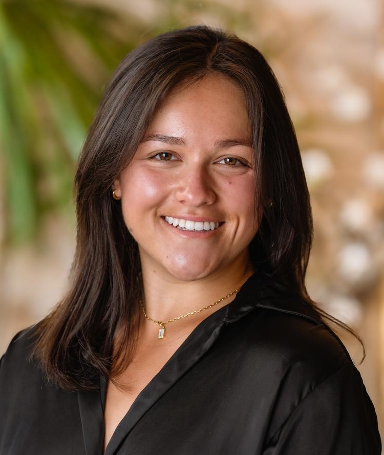 A woman with shoulder-length dark hair smiles while wearing a black button-up shirt and a gold necklace. She stands indoors with a blurred background featuring green plants and soft lighting.
