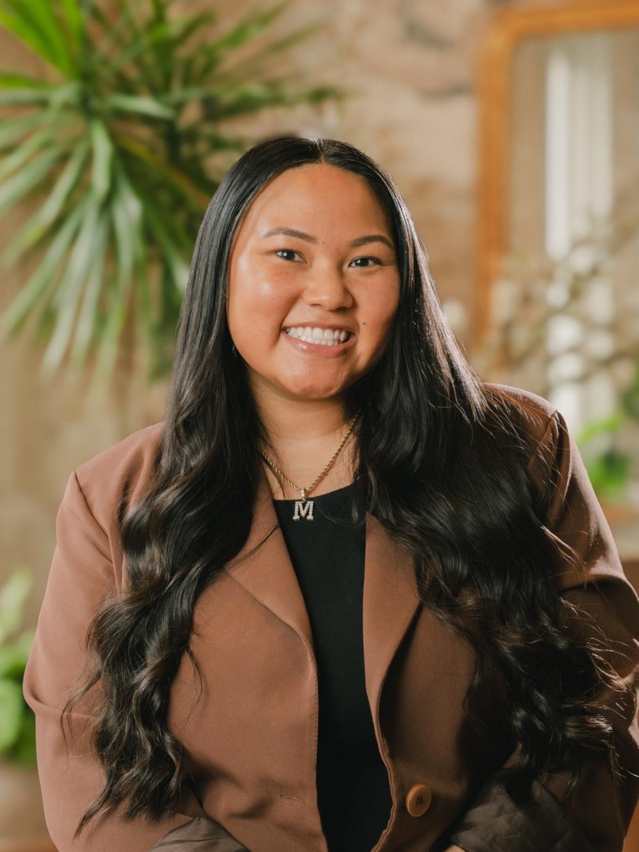 A woman with long, wavy black hair, wearing a brown blazer and a necklace with the letter M, smiles while sitting indoors with plants and a blurred mirror in the background.