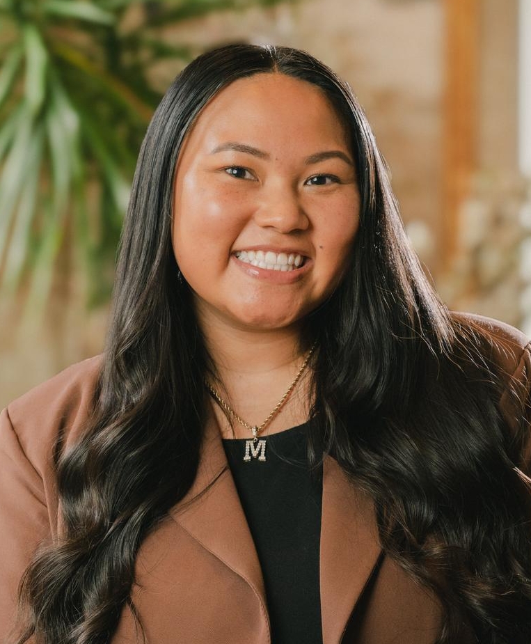 A woman with long, wavy black hair, wearing a brown blazer and a necklace with the letter M, smiles while sitting indoors with plants and a blurred mirror in the background.