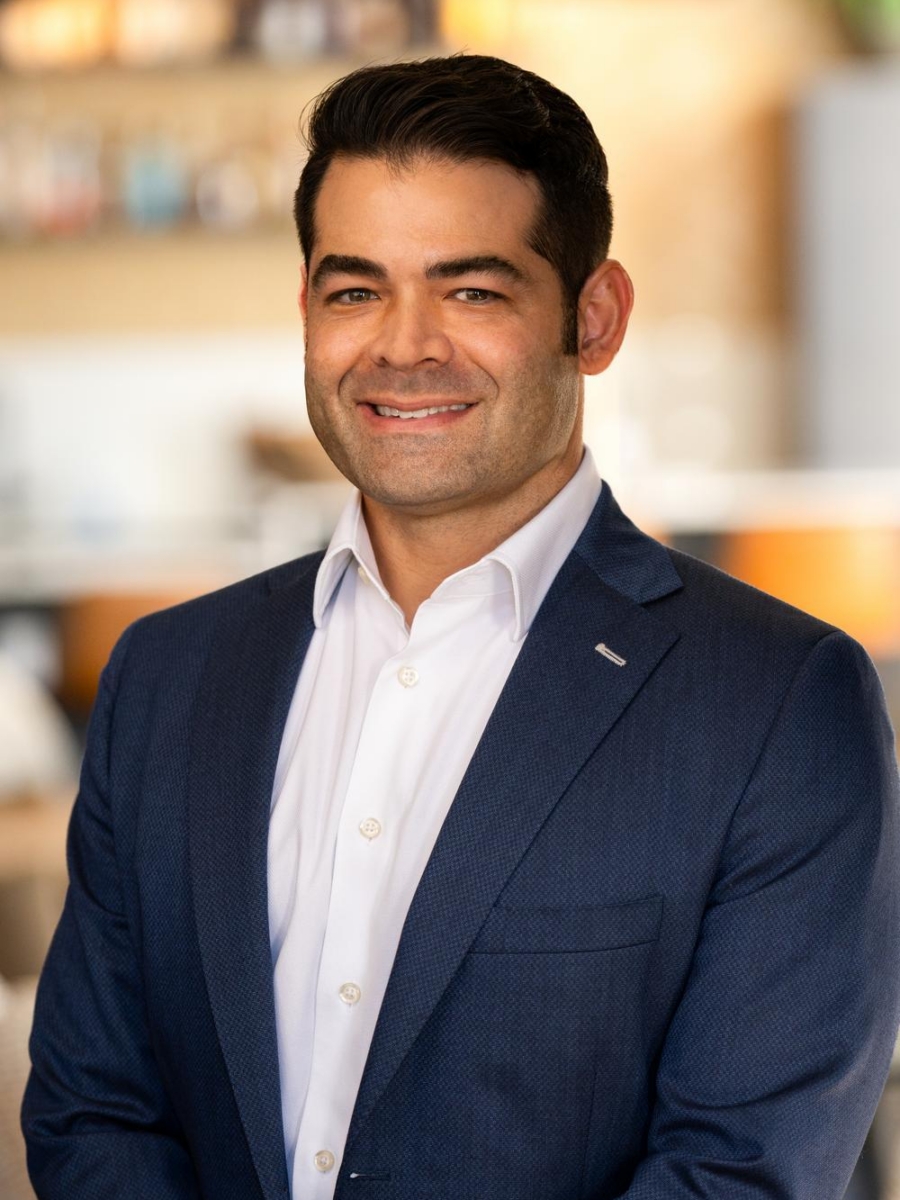 A man in a blue suit and white shirt smiles while standing indoors in a blurred, modern setting with warm lighting.
