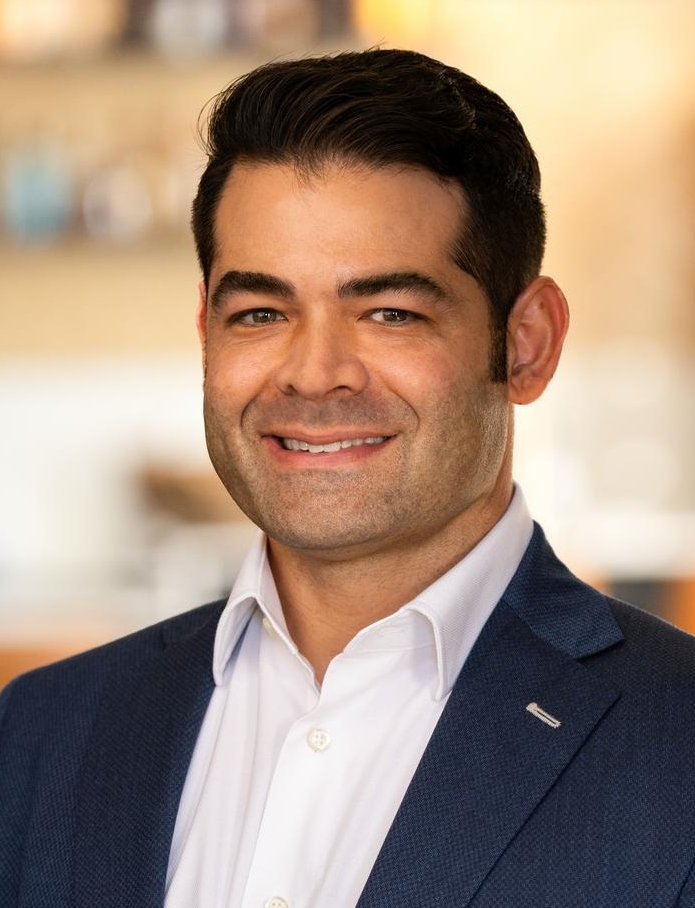 A man in a blue suit and white shirt smiles while standing indoors in a blurred, modern setting with warm lighting.