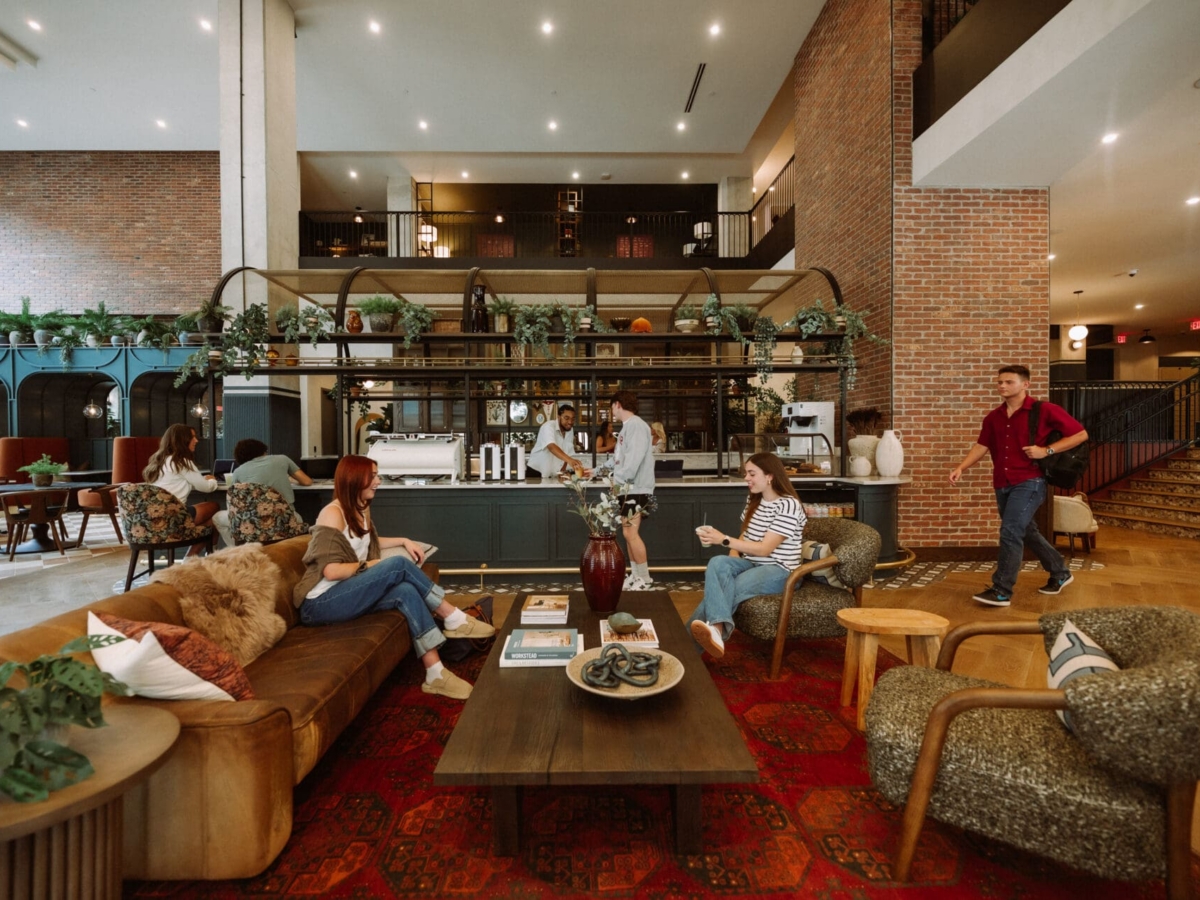A modern, spacious lounge at Rambler Columbus with people sitting on sofas and chairs, talking and working. A man prepares drinks at a counter, while others walk by. The room features warm lighting, plants, and brick walls near Ohio State University.