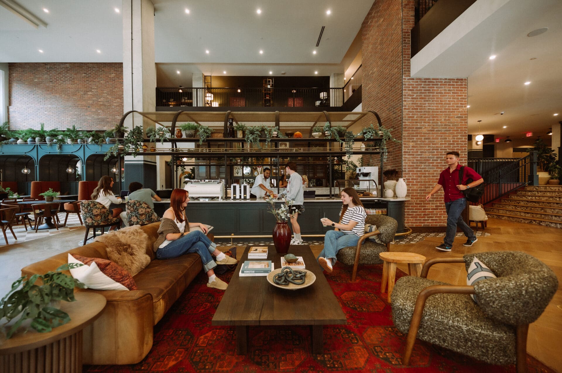 A modern, spacious lounge at Rambler Columbus with people sitting on sofas and chairs, talking and working. A man prepares drinks at a counter, while others walk by. The room features warm lighting, plants, and brick walls near Ohio State University.