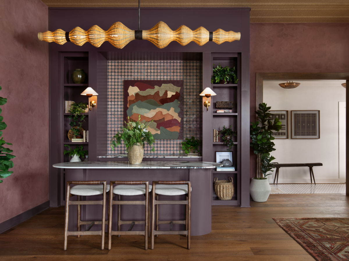 A stylish kitchen with a purple island, three stools, gold pendant light, abstract wall art, built-in shelves, and potted plants in a modern, earthy setting.